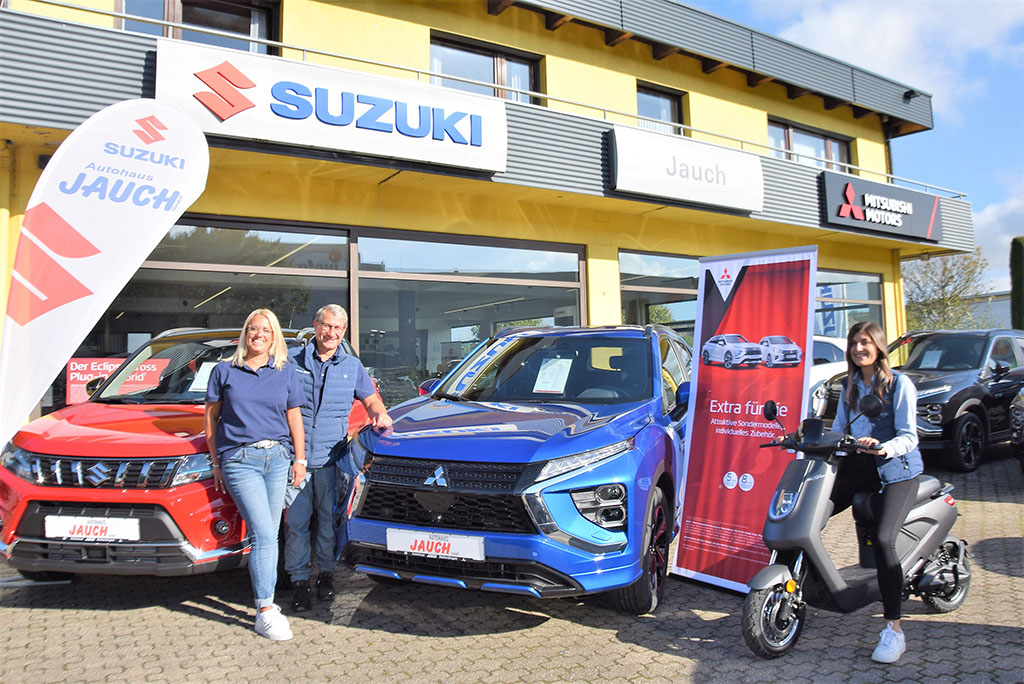 Familie Kübler, vor zwei Fahrzeugen links ein roter Suzuki und rechts ein blauer Mitsubishi sowie ein Roller im Hintergrund. Das Autohaus Gebäude