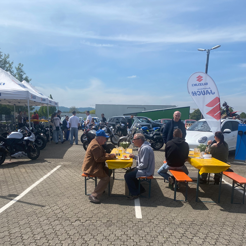 Von links nach rechts sehen wir aufgebaute Pavillon mit darunter stehenden Motorrädern eine Gruppe. Junge Menschen unterhält sich über die ausgestellten Fahrzeuge rechts davon sehen wir zwei Biertische mit verschiedenen Personen und rechts davon ein Job Fleck mit dem Logo Suzuki sowie Autohaus Jauch.