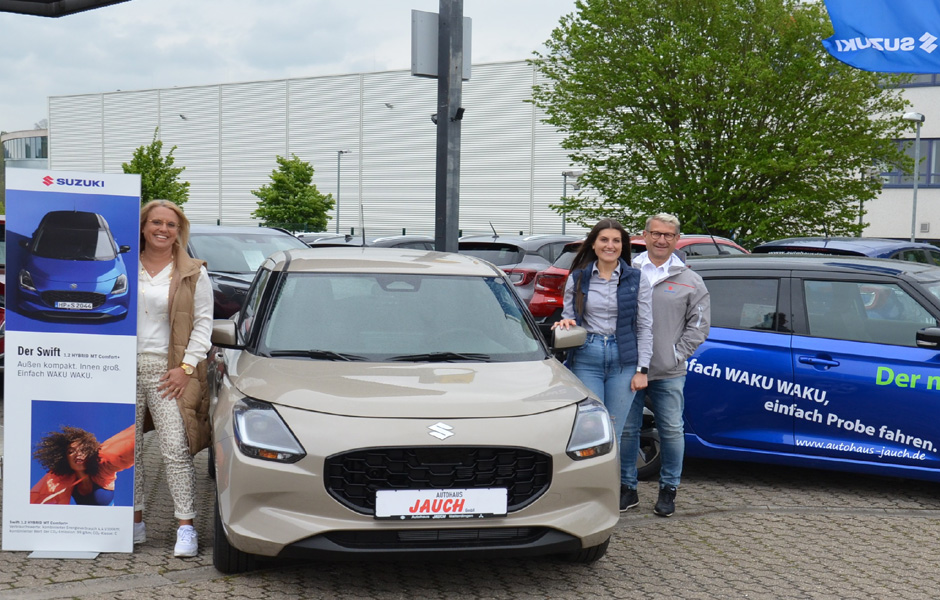 Wir sehen Frau Kübler mit einem präsenter links neben einem Suzuki in Beyer Farbe. Danach folgt Herr Kübler mit Tochter und ganz rechts ein blauer Suzuki im Hintergrund ein Baum sowie die Außenfassade der Nachbar Firma