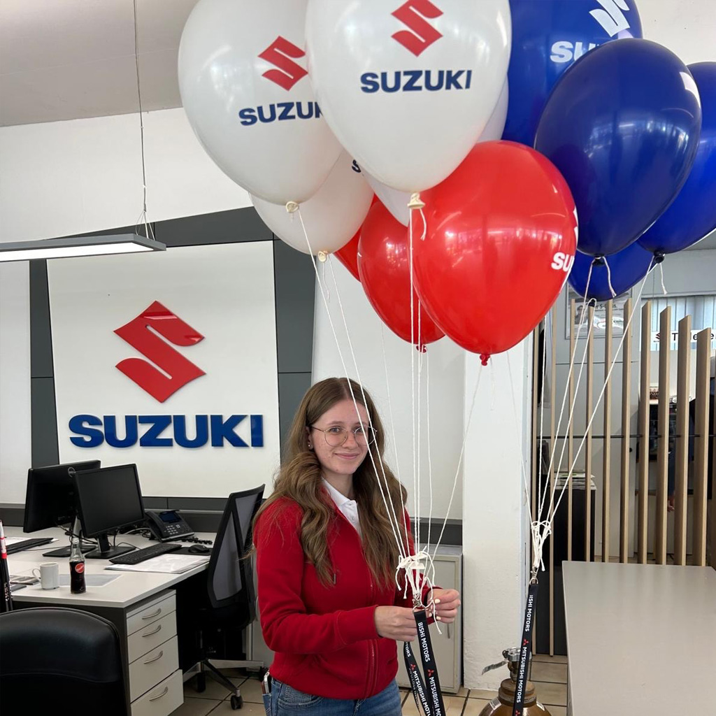 Wir sehen von links nach rechts, das Logo Suzuki rechts. Daneben steht eine junge Dame in rotem Sweat Oberteil mit verschiedenen Luftballons in den Farben. Weiß rot, blau.